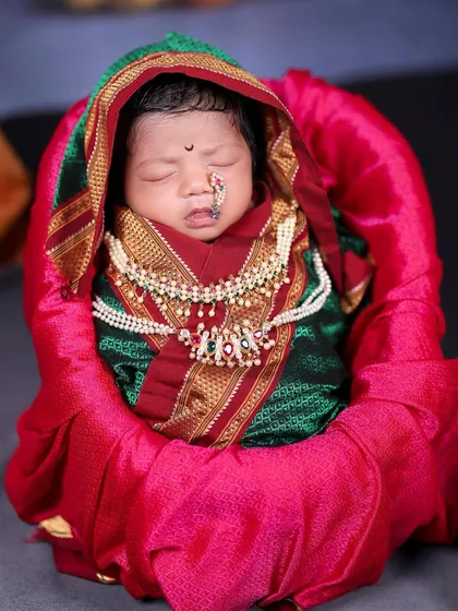 A close-up of a sleeping newborn dressed in a traditional green and red Maharashtrian saree, complete with a tiny nose ring (nath) and jewelry.