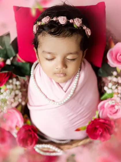 Pretty in pink. This newborn girl is wrapped snugly and adorned with a pearl necklace and a floral headband, surrounded by soft roses.
