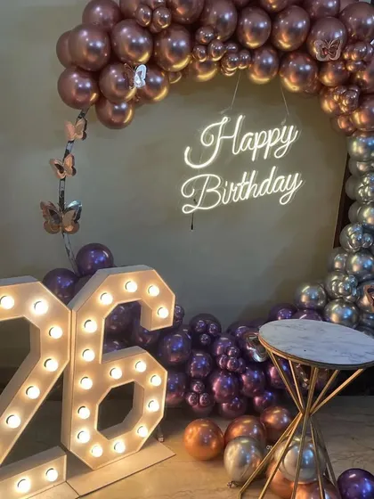 A beautiful two-tone balloon decoration for a 26th birthday. The arch combines metallic rose gold and purple balloons, with butterfly details, a neon sign, and large marquee numbers.