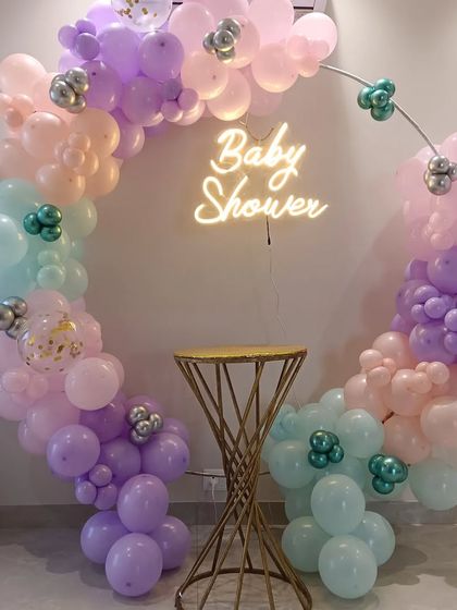 A chic and simple baby shower decoration. This setup features a circular metal frame adorned with a pastel rainbow balloon garland and a 'Baby Shower' neon sign, with a small table ready for a cake or gifts.