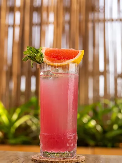 A refreshing pink grapefruit cocktail against a lush green, tropical background. The styling is simple and clean, focusing on the drink's vibrant colour and fresh garnish.