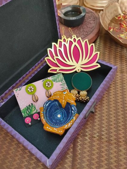 An affordable and beautiful Diwali gift box. This hamper includes a hand-painted diya, two pairs of earrings, and a decorative lotus wall hanging.