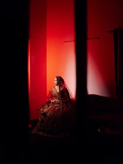 An artistic and moody bridal portrait. The bride is seated in a dark room, her silhouette illuminated by a dramatic red light, creating a powerful and cinematic feel.
