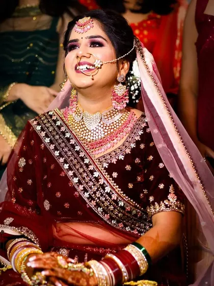 The pure joy of a bride on her wedding day. This candid photo captures a moment of genuine laughter, showcasing her beautiful smile. The warm lighting enhances the rich maroon color of her velvet lehenga and the sparkle of her elaborate pink and white jewelry set.