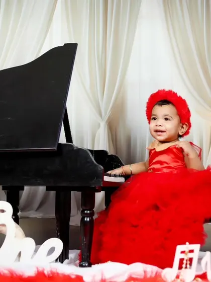 A one-year-old girl in a festive red dress plays the piano to celebrate her birthday.