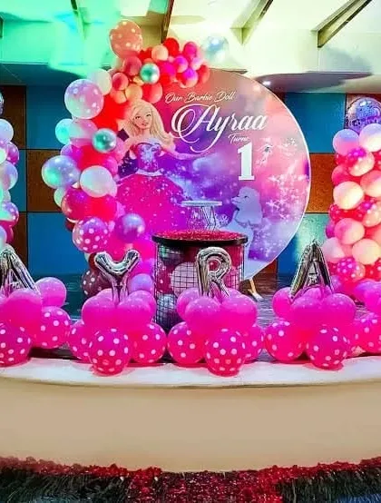 A Barbie-themed first birthday party on a stage. This decoration features a "Barbie Doll" backdrop, balloon columns, and hot pink balloon arrangements.