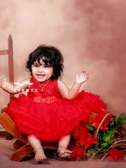 A joyful portrait of a one-year-old baby girl in a vibrant red dress. The simple, warm-toned backdrop allows her happy expression and beautiful outfit to be the main focus of the photograph.