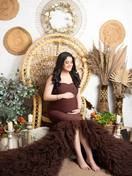 A moment of quiet reflection in our bohemian-rustic set. The expectant mother is seated on a peacock chair, wearing a beautiful brown gown with a ruffled train. The earthy tones and natural textures create a warm and tranquil atmosphere.