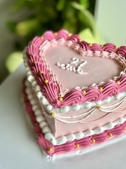A close-up of a pink heart-shaped vintage cake, showing the detailed layers of piping in different shades of pink and white, accented with tiny gold beads.
