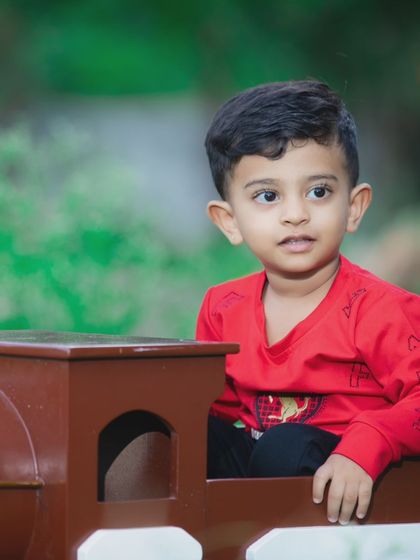 A candid shot of a little boy in a red shirt, sitting inside a toy train prop during an outdoor session.