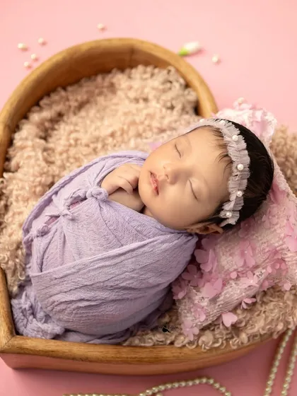 A peaceful newborn girl swaddled in lavender, resting in a heart-shaped bowl. The pearls add a touch of elegance to this classic newborn photo.