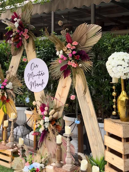 We love creating unique, boho-chic setups for life's special moments. This baby shower backdrop features A-frame wood structures adorned with dried palms, pampas grass, and vibrant floral arrangements for a warm, rustic feel.
