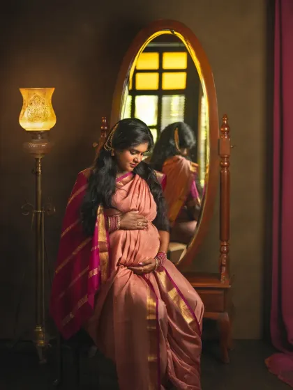 A beautiful studio portrait of an expecting mother in a traditional silk saree, seated before a vintage mirror. The warm light from the lamp adds a golden glow, creating a rich, classic composition.