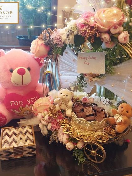 A delightful Teddy Day gift hamper combining sweet treats with a cuddly companion. This arrangement includes a variety of our handmade chocolates, a miniature cart, and a soft teddy bear, all presented in a decorative floral hoop.