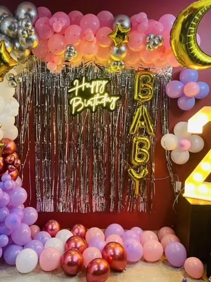 A fun and festive birthday decoration with a silver fringe curtain and a colorful balloon arch. The setup includes a light-up number '2' and 'BABY' foil balloons.