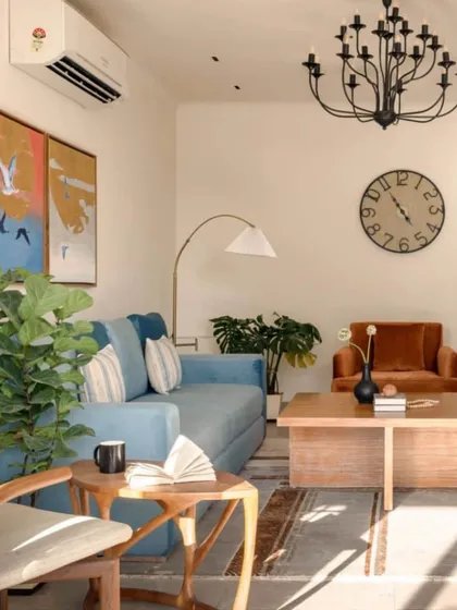 The living room is bathed in natural light, highlighting the mix of colors and textures. The space is anchored by a large clock and a classic chandelier, blending modern and traditional elements.