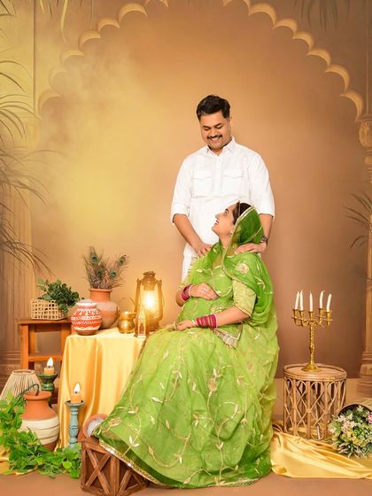 A joyful couple's portrait celebrating their heritage. The mom-to-be is seated, wearing a beautiful green traditional outfit, while her partner stands lovingly behind her in our rustic, warm-toned set.