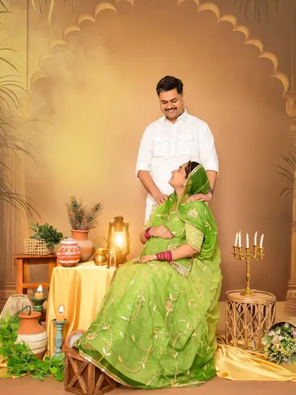 A joyful couple's portrait celebrating their heritage. The mom-to-be is seated, wearing a beautiful green traditional outfit, while her partner stands lovingly behind her in our rustic, warm-toned set.