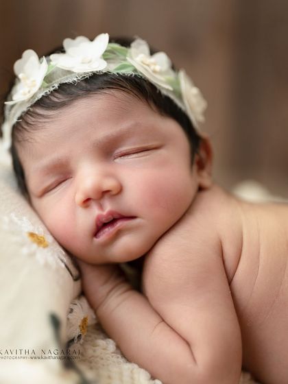 A one-week-old baby girl sleeping on a tiny pillow, wearing a delicate white floral headband. The rustic wooden background adds warmth and texture to this classic portrait.