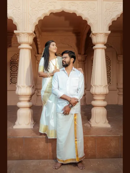 A classic and regal portrait from a South Indian themed pre-wedding shoot. The couple's coordinated white and gold traditional outfits look stunning against the carved stone pillars.