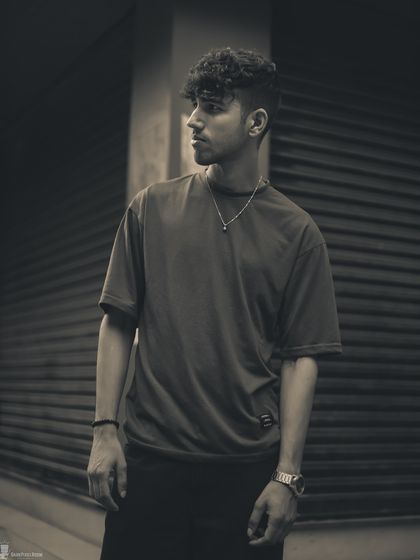 A moody, sepia-toned black and white portrait of a male subject. The urban background and strong pose create a classic, masculine image with a timeless quality.