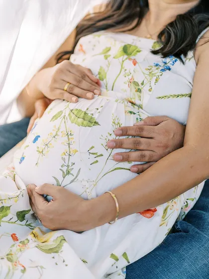 A close-up detail shot of the couple's hands embracing the baby bump. Her floral dress perfectly complements the natural, outdoor setting of the photoshoot.