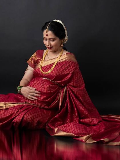 A classic pose in a rich red silk saree against a dark backdrop. The reflection on the floor adds a touch of modern studio elegance to this traditional maternity portrait.