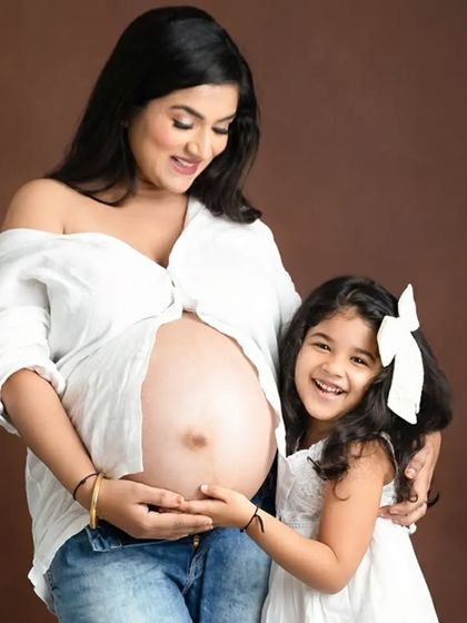 A joyful family moment captured against a warm brown backdrop. The daughter's happy expression as she touches her mother's baby bump makes this a precious memory.