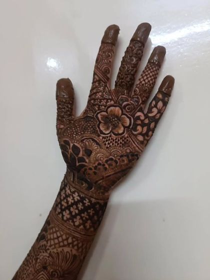 A close-up of a bridal hand with a central flower and detailed patterns covering the fingers and wrist. The fingertips are covered in henna for a deep, traditional stain.