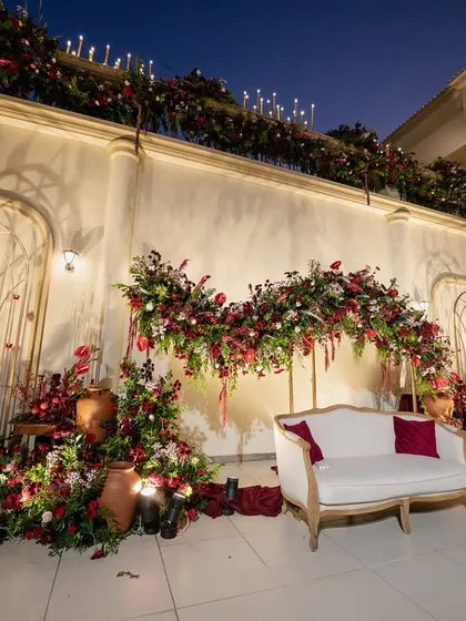 A twilight view of a regal reception stage. The warm lighting and deep red florals create a dramatic and romantic ambiance against the evening sky.