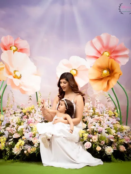 A mother and daughter share a sweet moment sitting together in a dreamy, floral wonderland. The soft, pastel colors and their gentle interaction create a tender and artistic portrait.