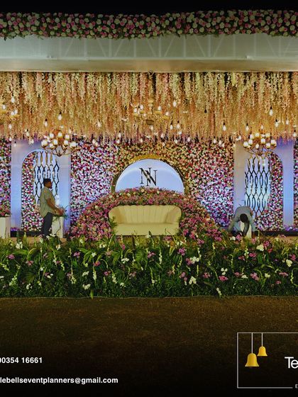 A grand reception stage featuring a massive floral wall, a custom couple's monogram, and a ceiling dripping with wisteria and Edison bulbs for a magical glow.