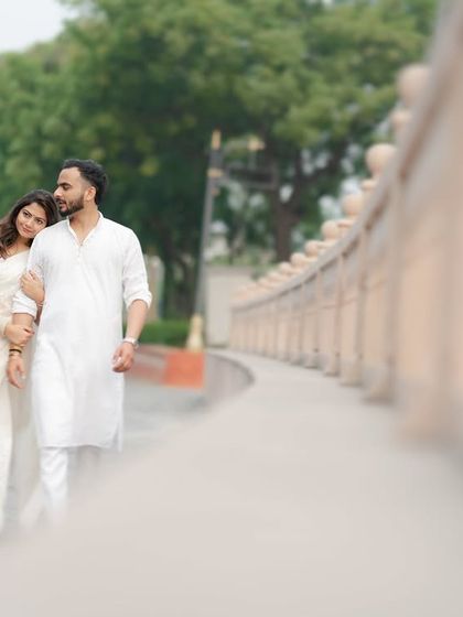 A serene and romantic pre-wedding photo of a couple dressed in white, walking together along a stone causeway. The soft focus and natural setting create a dreamy, peaceful atmosphere.
