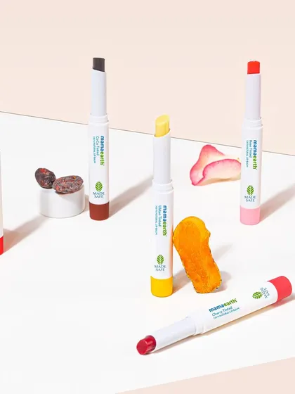 A group shot of Mamaearth's tinted lip balms, styled with their corresponding natural ingredients like orange, rose, and cocoa. The clean, geometric arrangement makes the product line easy to understand at a glance.