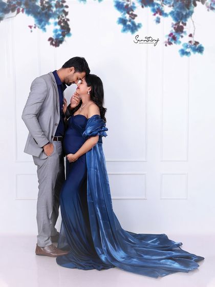 An intimate couple portrait against a clean white background. The husband's tender embrace and the wife's elegant blue gown create a timeless and romantic image.