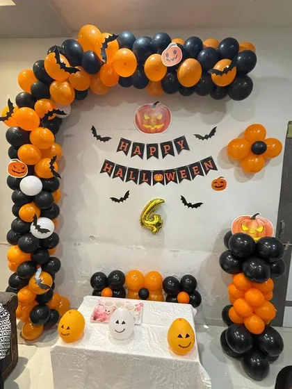 A fun and spooky Halloween party decoration for a home celebration. The setup features a black and orange balloon arch, a 'Happy Halloween' banner, bat cutouts, and themed balloons shaped like ghosts and pumpkins to create the perfect party backdrop.