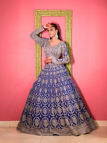 A full-length shot against a vibrant pink wall, making the blue of the lehenga pop.