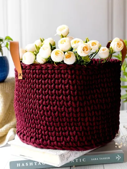 This rich, burgundy-colored crochet basket is filled with white flowers, creating a beautiful contrast. Styled on a stack of books, it's shown as a versatile and elegant decor piece.