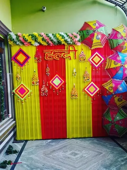 Here is a classic Haldi ceremony backdrop I put together. The combination of bright red and yellow drapes, traditional hangings, and colorful Rajasthani umbrellas creates a festive and picture-perfect setting for the event.