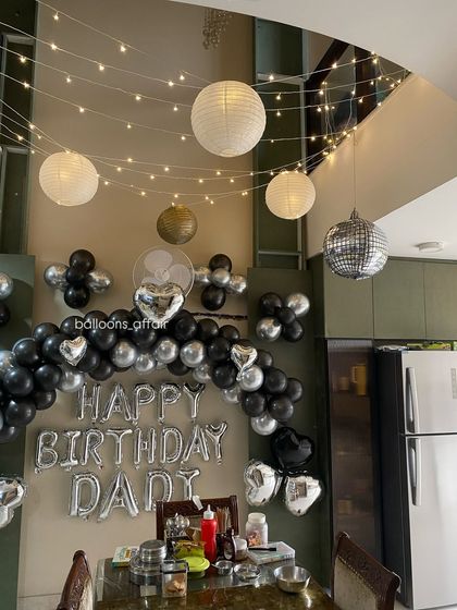 A cozy 'Happy Birthday Dad' decoration in a dining area. The setup features a black and silver balloon arch, hanging lanterns, and fairy lights.