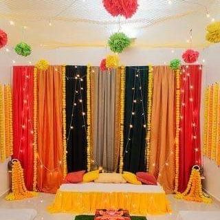 A colorful indoor setup with drapes in orange, black, and red, decorated with marigold garlands and fairy lights. This design is perfect for a home-based Mehandi or Sangeet function.