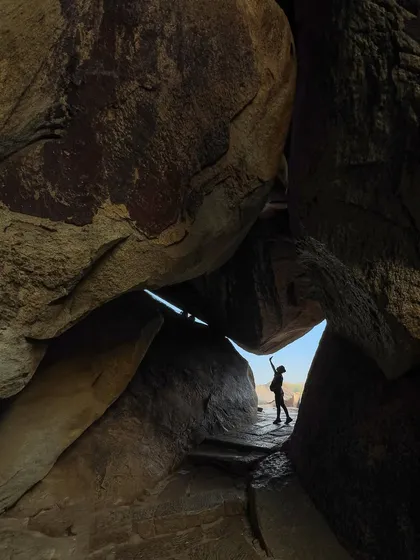 A traveler reaches towards the sky from within a rock cave in Hampi. This shot uses silhouette and natural framing to convey a sense of scale, wonder, and adventure in a historic landscape.