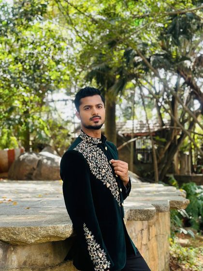 Posing in a lush, natural setting wearing a dark green embroidered bandhgala. The contrast between the formal wear and the rustic background creates a striking image.