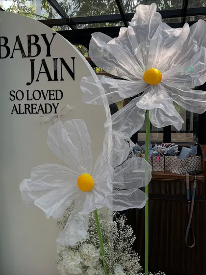 The whimsical, larger-than-life daisies stand next to the 'Baby Jain So Loved Already' sign, adding a touch of playful sophistication.
