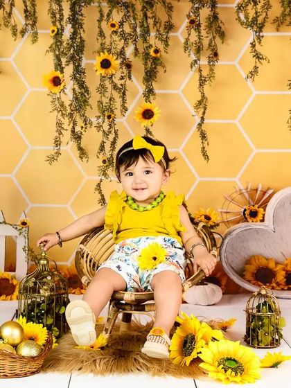 A baby girl sits on a miniature chair in a cheerful sunflower and honeycomb-themed setup. A perfect theme for a sunny personality.