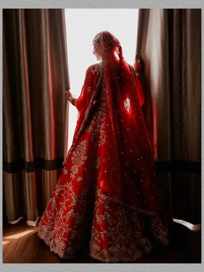 A beautiful silhouette of the bride looking out a window. This artistic shot highlights the stunning details of her red lehenga and veil against the soft light.