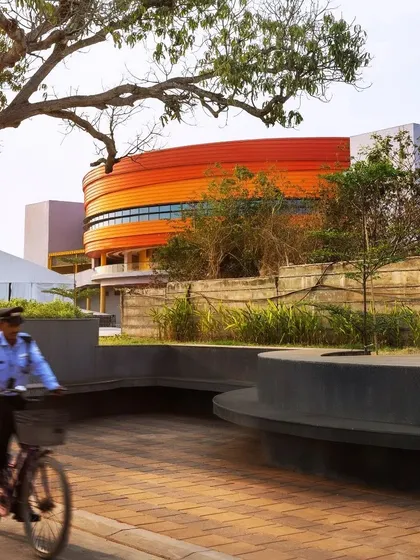The MARC Auditorium at the MAHE Bengaluru Campus, seen from a landscaped plaza. The building's curved orange form creates a visual anchor, while the surrounding public spaces invite community use.