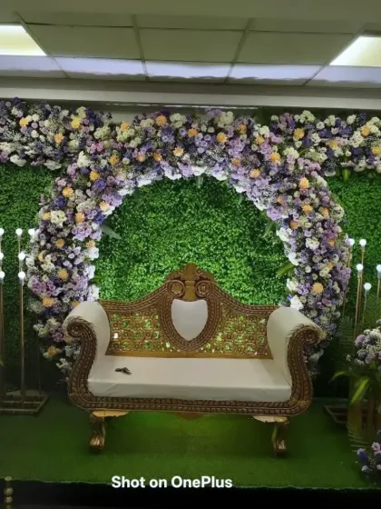 A lush green backdrop for a Half-Saree function in JP Nagar. The artificial green wall is adorned with a thick arch of pastel purple and white flowers, creating a garden-like feel indoors.