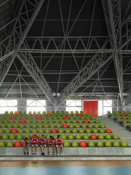 A group of athletes rests on the tiered seating inside the sports arena. The vibrant green and orange seats add a splash of color to the otherwise neutral interior palette of concrete and steel.