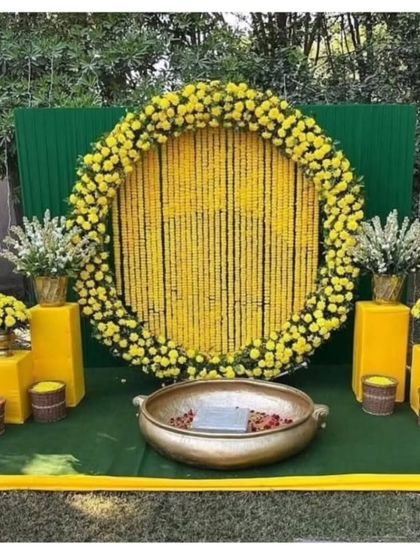 A classic Haldi backdrop for outdoor functions. This setup features a large circular frame filled with strings of yellow marigolds, a green panel background, and a traditional brass urli in the center for the ceremony.